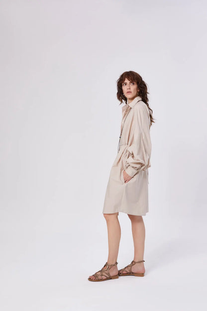 Eine Frau mit gewelltem braunem Haar steht seitlich vor einem unifarbenen Hintergrund und trägt das lockere, langärmelige Antonia Dress Vol. 2 Beige mit Taschen und braune Riemchensandalen.