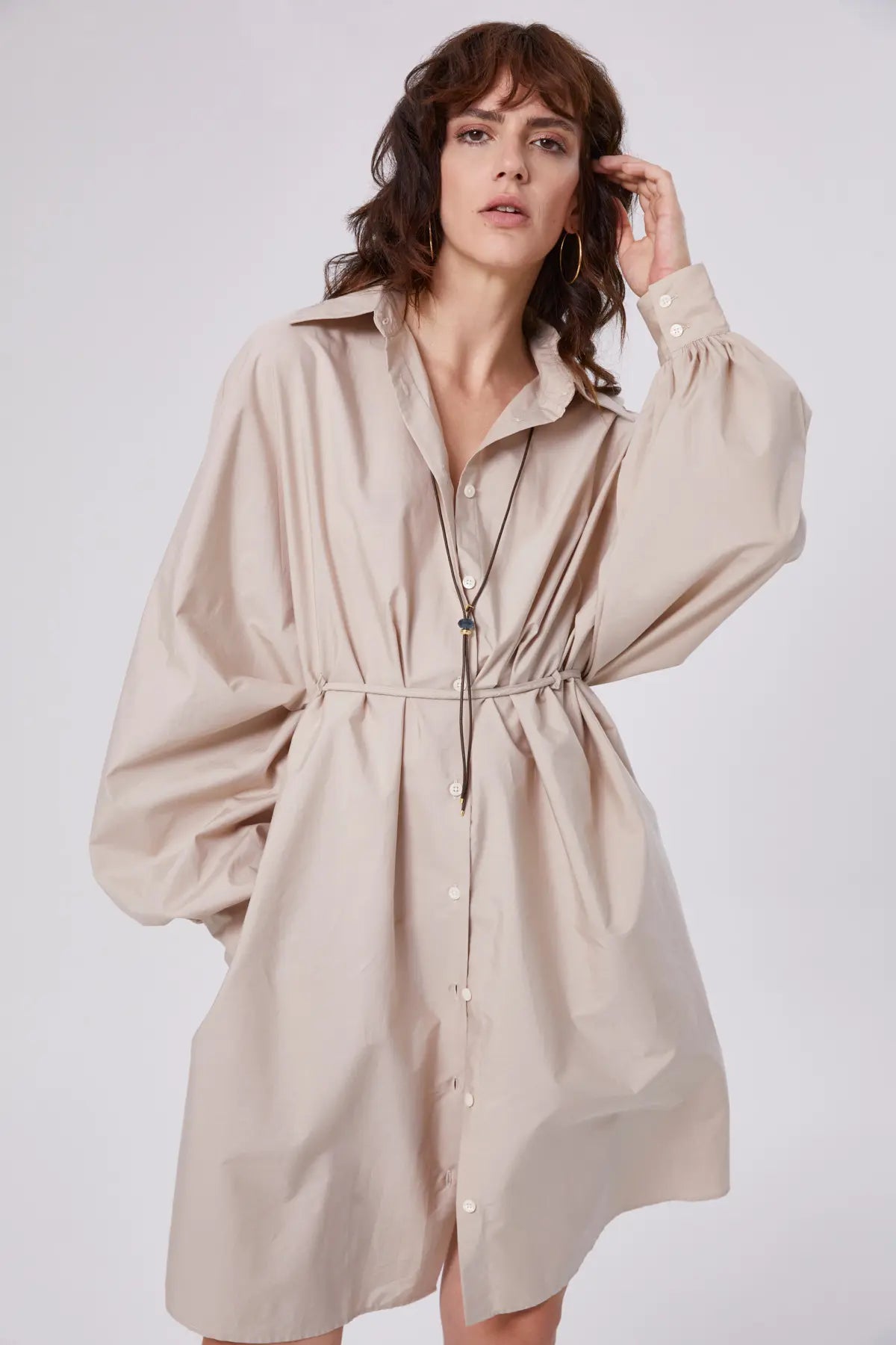 In dem übergroßen Kleid Antonia Dress Vol. 2 Beige mit langen Ballonärmeln und einem dünnen Taillenband steht eine Person vor einem schlichten Hintergrund und blickt selbstbewusst in die Kamera.
