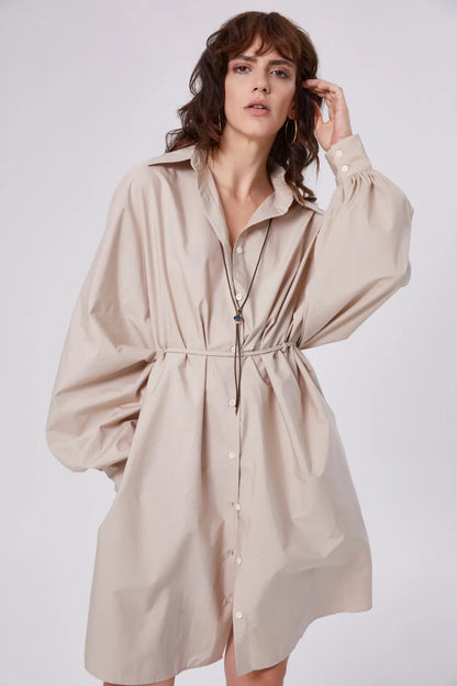 In dem übergroßen Kleid Antonia Dress Vol. 2 Beige mit langen Ballonärmeln und einem dünnen Taillenband steht eine Person vor einem schlichten Hintergrund und blickt selbstbewusst in die Kamera.