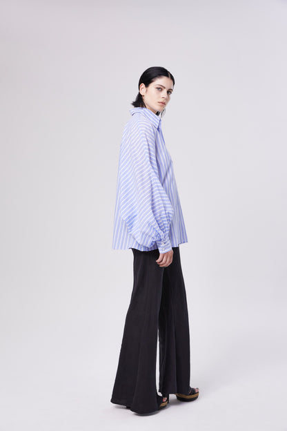 Eine Person mit glattem dunklem Haar, die den blauen Polo Antonia-Streifen und eine schwarze Hose mit weitem Bein trägt, steht seitlich vor einem schlichten, hellen Hintergrund.