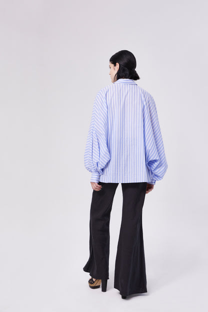 Eine Person mit kurzen dunklen Haaren, von hinten gesehen, trägt einen blauen Polo Antonia-Streifen und eine schwarze Schlaghose und steht vor einem schlichten hellen Hintergrund.