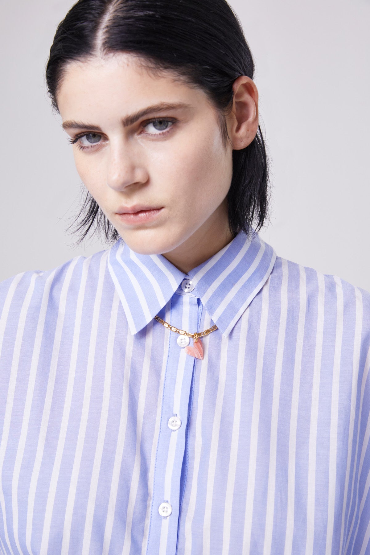 Eine Person mit glattem schwarzem Haar, die das blau gestreifte Polo Antonia und eine goldene Halskette mit rosafarbenen und weißen Anhängern trägt, blickt mit neutraler Miene in die Kamera.