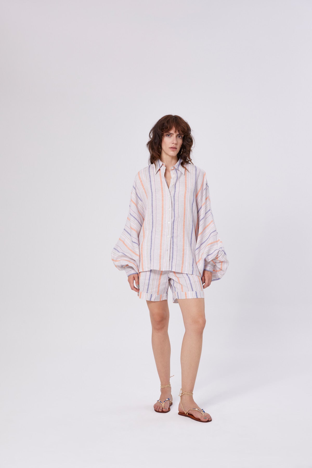Eine Frau steht vor einem schlichten weißen Hintergrund und trägt ein lockeres, langärmeliges Button-up-Hemd und dazu passende Shorts Linen Stripes Blue Orange in Pastelltönen, gepaart mit braunen flachen Sandalen.