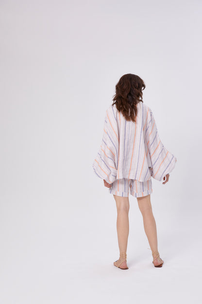 Eine Person mit gewelltem braunem Haar, von hinten gesehen in einem lockeren langärmeligen Oberteil und Shorts Leinenstreifen Blau Orange, kombiniert den Look mit flachen Sandalen vor einem schlichten weißen Hintergrund.