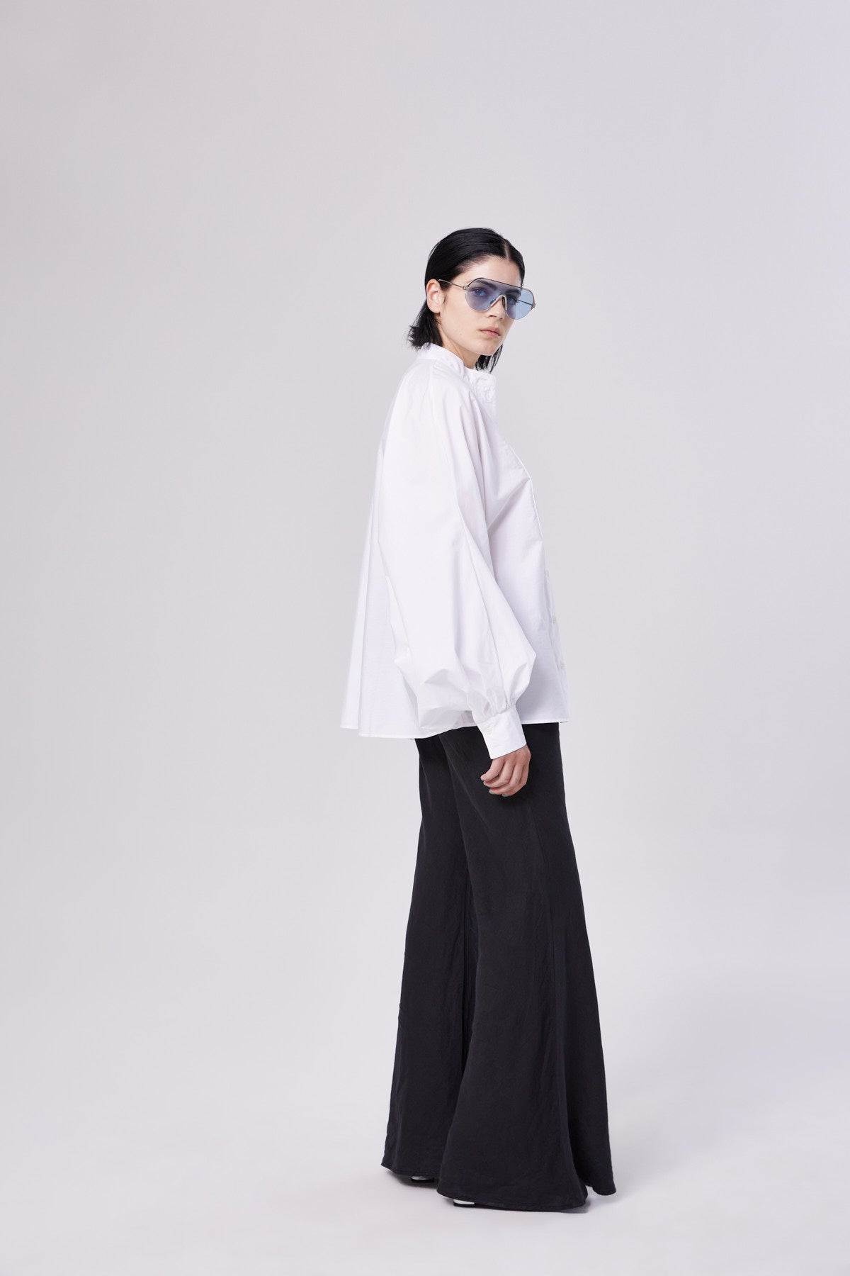 Eine Person mit zurückgebundenen dunklen Haaren trägt eine weite weiße Bluse, eine schwarze Joana-Hose mit ausgestellten Beinen und eine große blaue Sonnenbrille. Sie steht vor einem schlichten hellen Hintergrund und blickt in die Kamera.