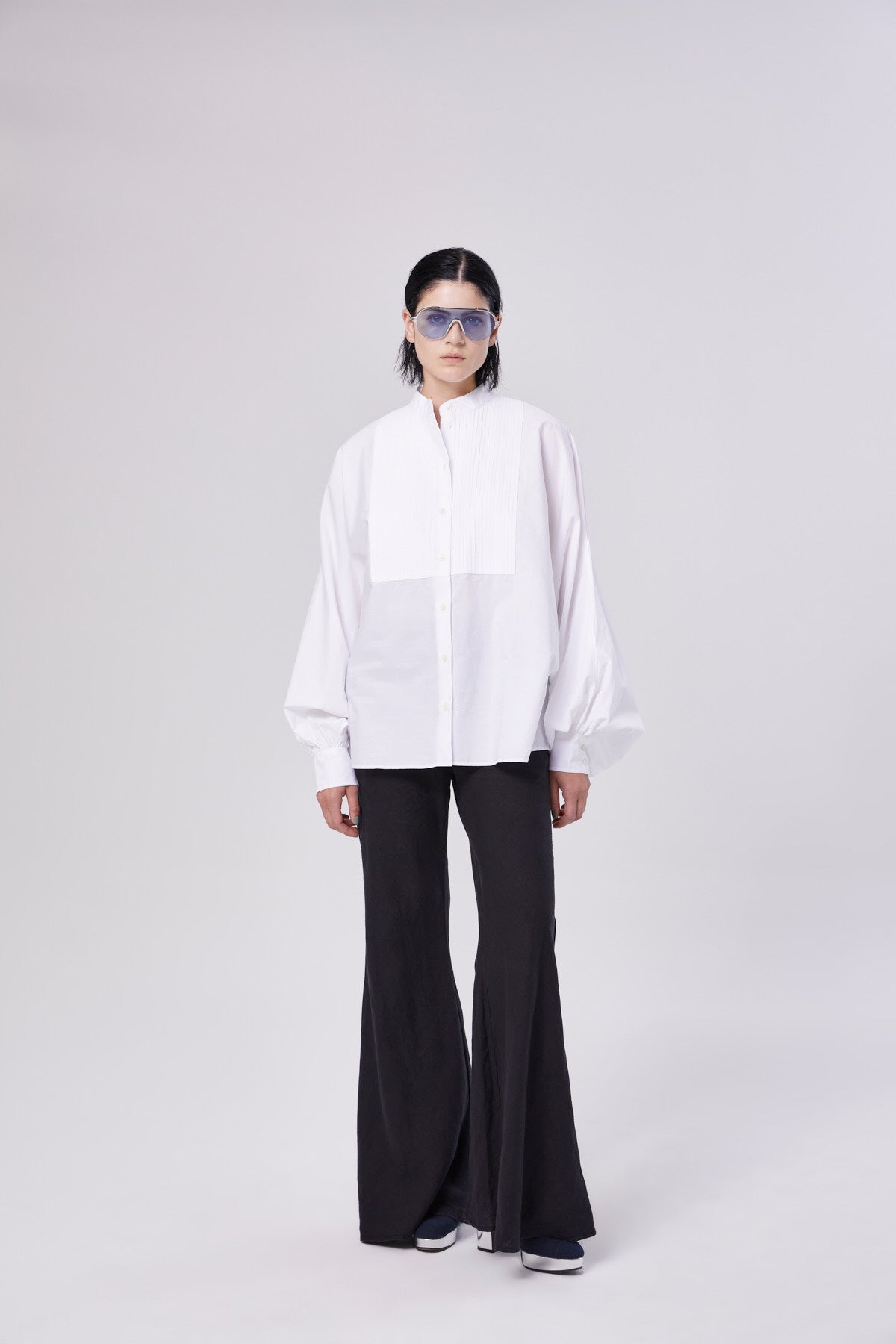 Eine Person trägt eine blaue Sonnenbrille, ein lockeres weißes Button-up-Hemd und die Hose Joana Black mit ausgestellten Beinen und steht vor einem schlichten hellen Hintergrund.