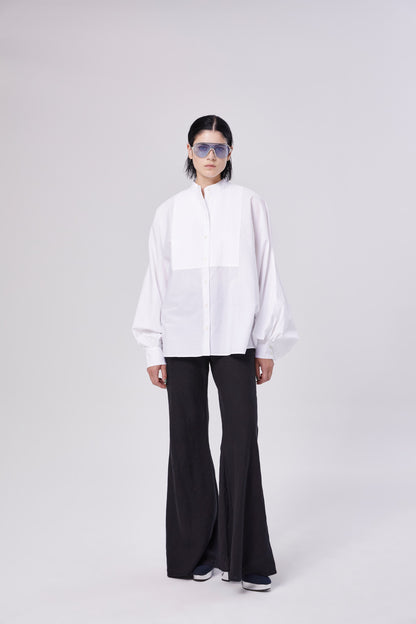 Eine Person trägt eine blaue Sonnenbrille, ein lockeres weißes Button-up-Hemd und die Hose Joana Black mit ausgestellten Beinen und steht vor einem schlichten hellen Hintergrund.