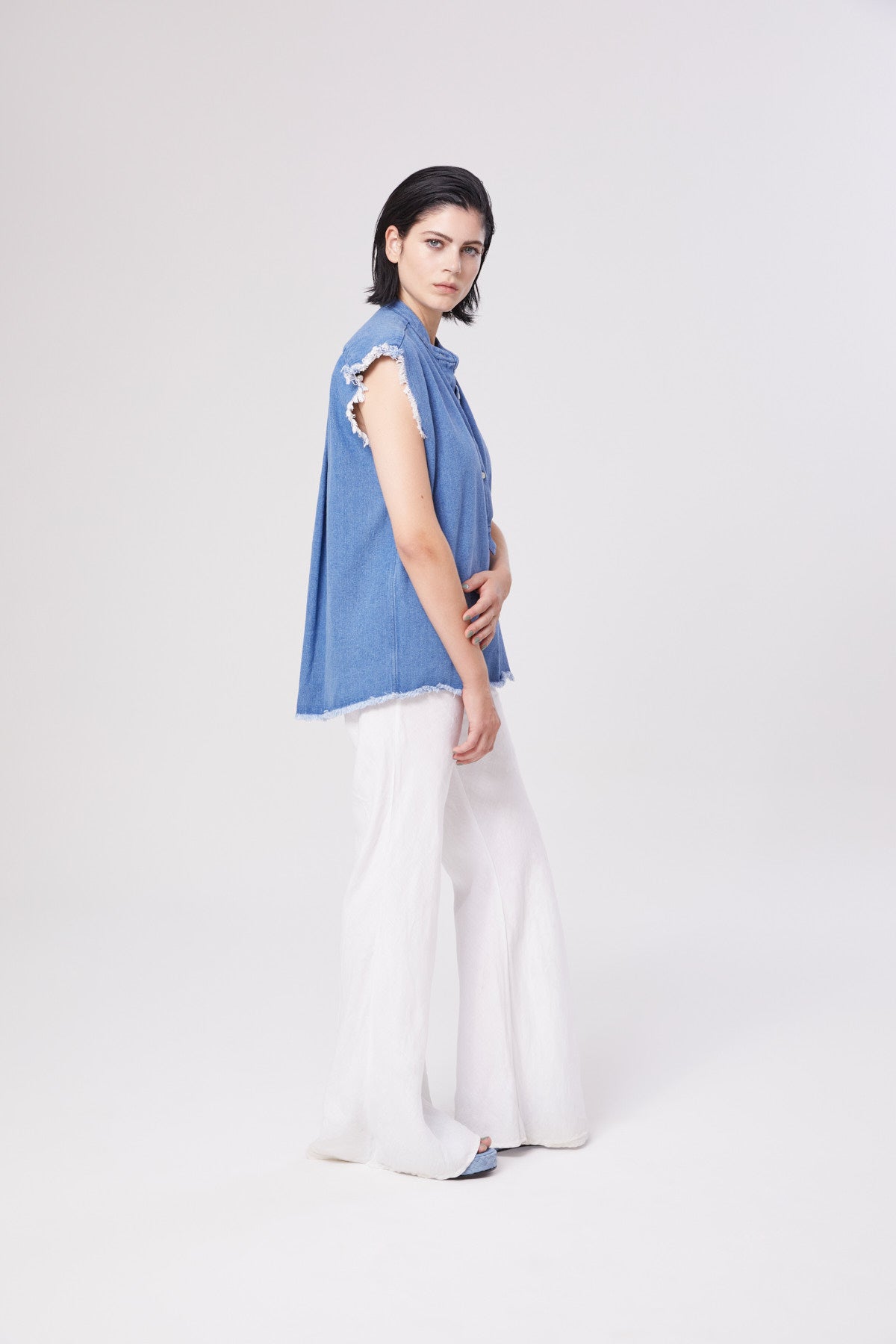 Eine Frau mit dunklem Haar steht seitlich und trägt die ärmellose blaue Weste mit ausgefransten Rändern von Barcelona Denim Vol 2 und eine weiße Hose mit weitem Bein über einer hellen Sommerbluse Damen vor einem schlichten hellen Hintergrund.