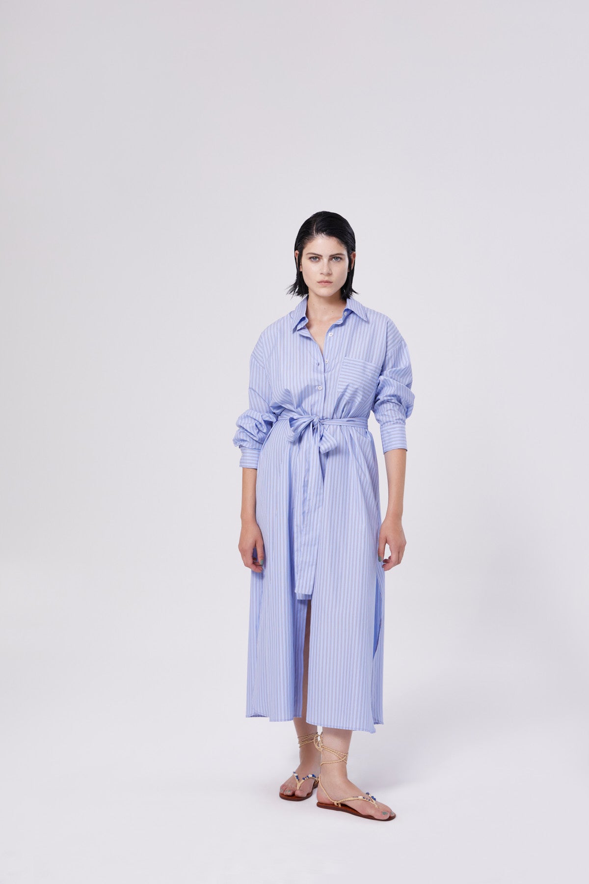 Eine Frau mit glattem dunklem Haar und neutralem Gesichtsausdruck trägt das Dress Scandi, ein langes blau-grau gestreiftes Hemdkleid mit gekrempelten Ärmeln, das in der Taille gebunden wird, sowie beigefarbene Sandalen und steht vor einem schlichten weißen Hintergrund.