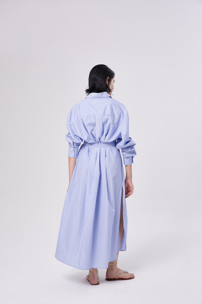 Eine Person mit kurzen dunklen Haaren steht mit dem Gesicht zur Wand und trägt das "Dress Scandi", ein langes, blau-grau gestreiftes Frauenkleid mit Gürtel und Seitenschlitzen, kombiniert mit flachen Sandalen vor einem einfachen, hellen Hintergrund.