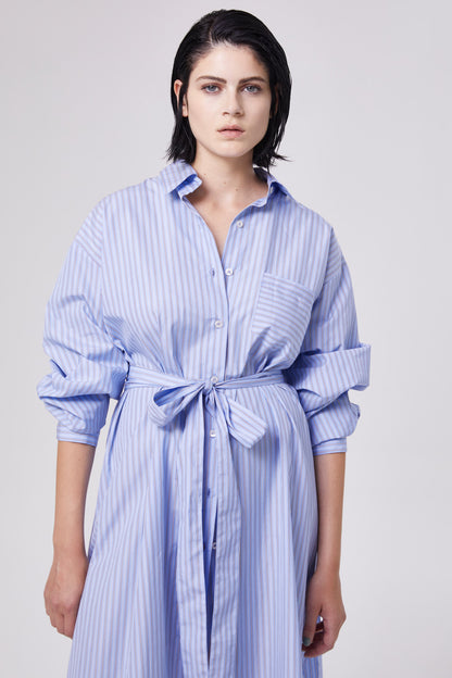 Eine Person mit kurzen dunklen Haaren trägt das Dress Scandi, ein blau-grau gestreiftes, gegürtetes Kleid mit hochgekrempelten Ärmeln, steht vor einem schlichten hellen Hintergrund und schaut mit neutralem Blick direkt in die Kamera.