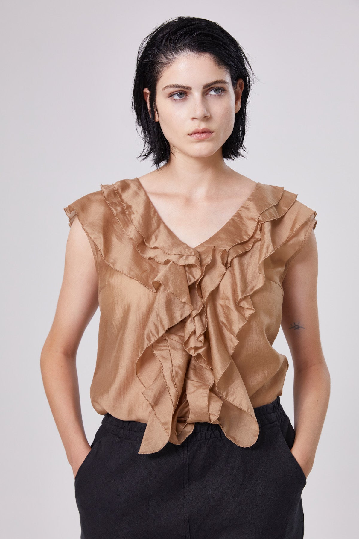 Eine Person mit kurzen dunklen Haaren trägt ein Top Ruffle Gold und eine schwarze Hose und steht mit den Händen in den Taschen vor einem schlichten hellen Hintergrund.