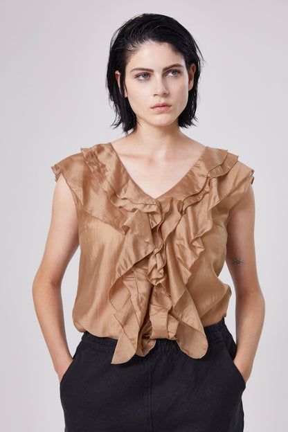 Eine Person mit kurzen dunklen Haaren trägt ein Top Ruffle Gold und eine schwarze Hose und steht mit den Händen in den Taschen vor einem schlichten hellen Hintergrund.