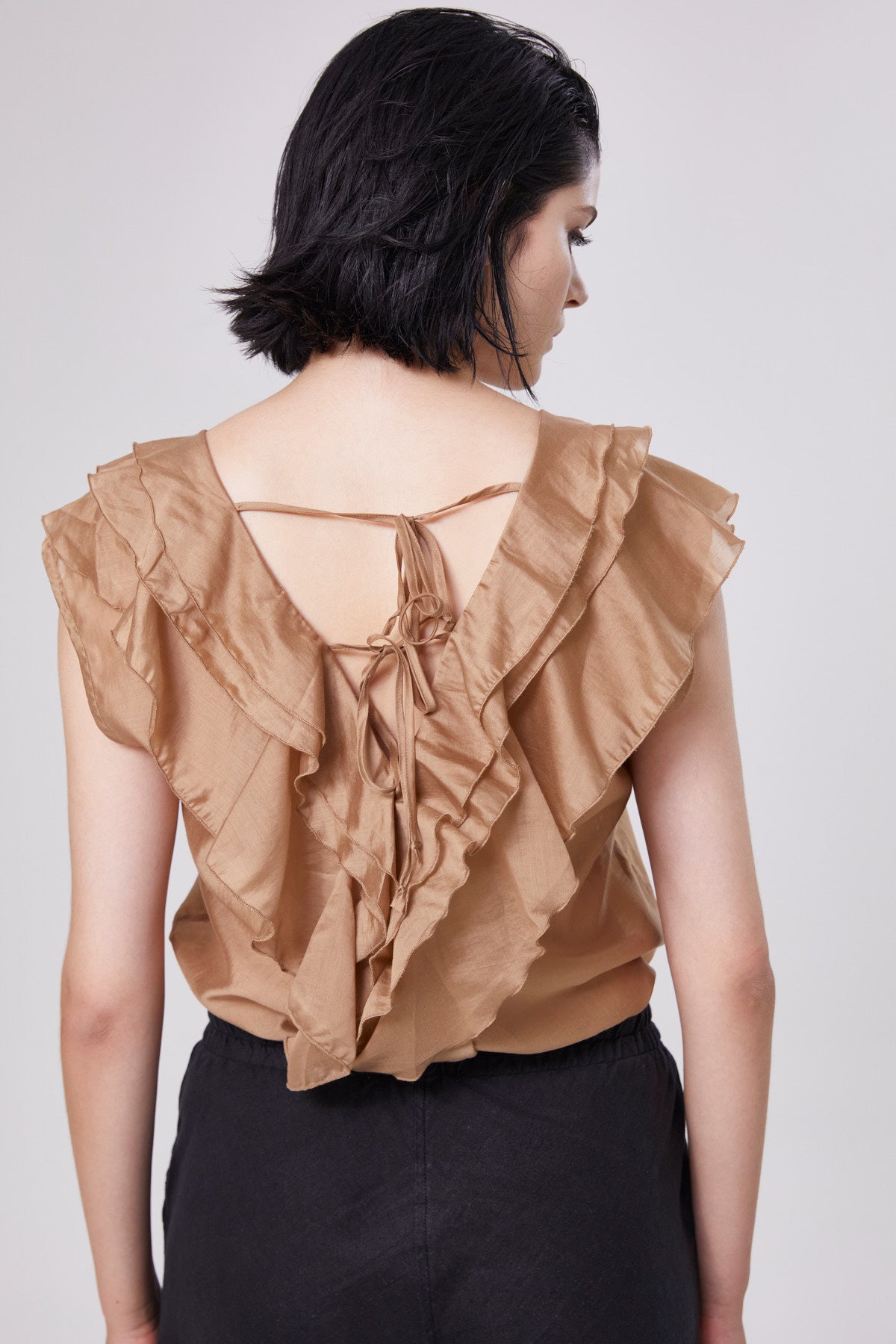 Eine Frau mit kurzen schwarzen Haaren ist von hinten zu sehen, wie sie das ärmellose Top Ruffle Gold mit einer Fliege auf dem Rücken trägt, gepaart mit einer schwarzen Hose mit hoher Taille für einen eleganten Look.