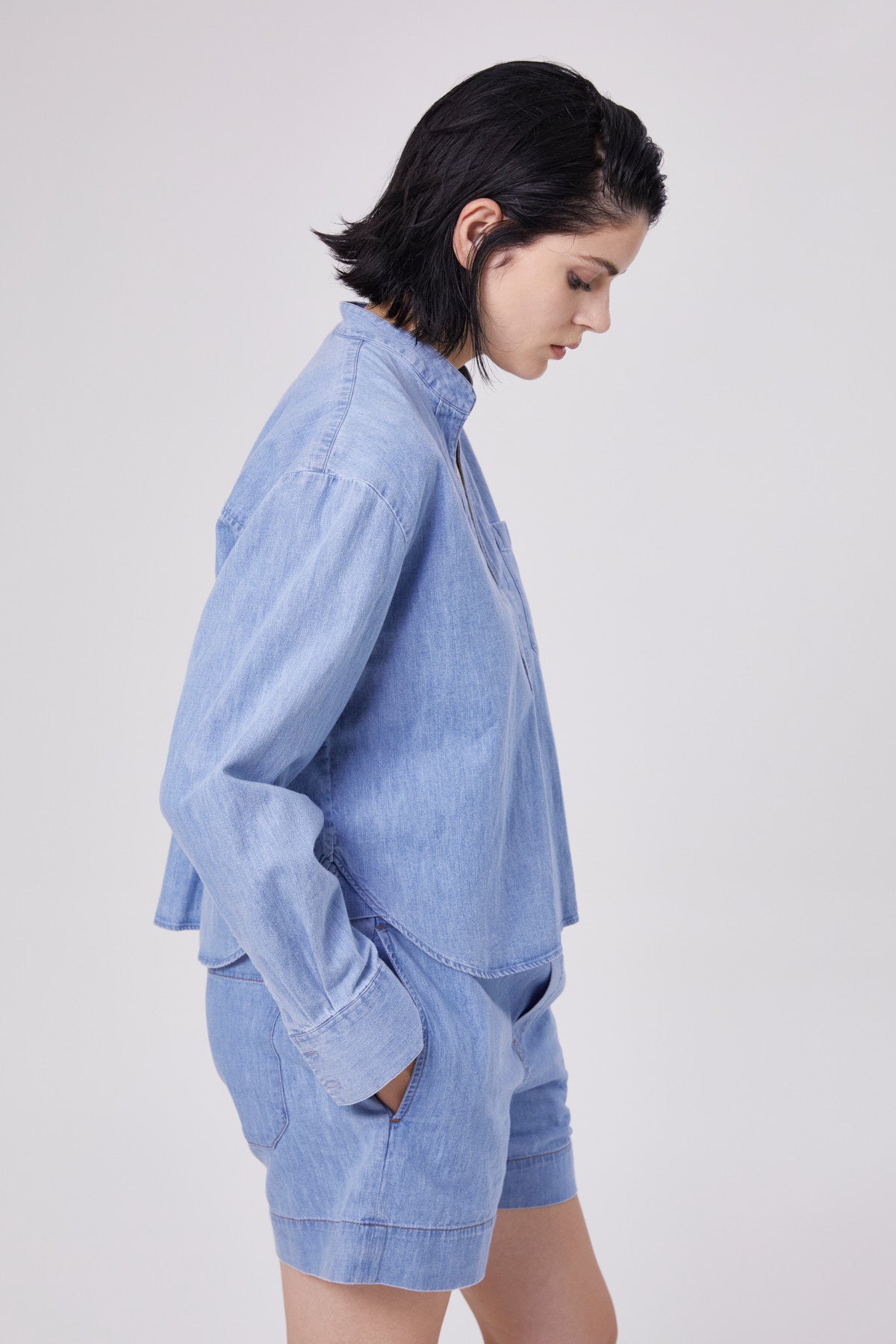 Eine Frau mit kurzen dunklen Haaren steht im Profil vor einem schlichten hellen Hintergrund. Sie trägt eine übergroße Bluse Polo Denim und dazu passende Jeans-Shorts, die Hände in den Taschen versteckt.