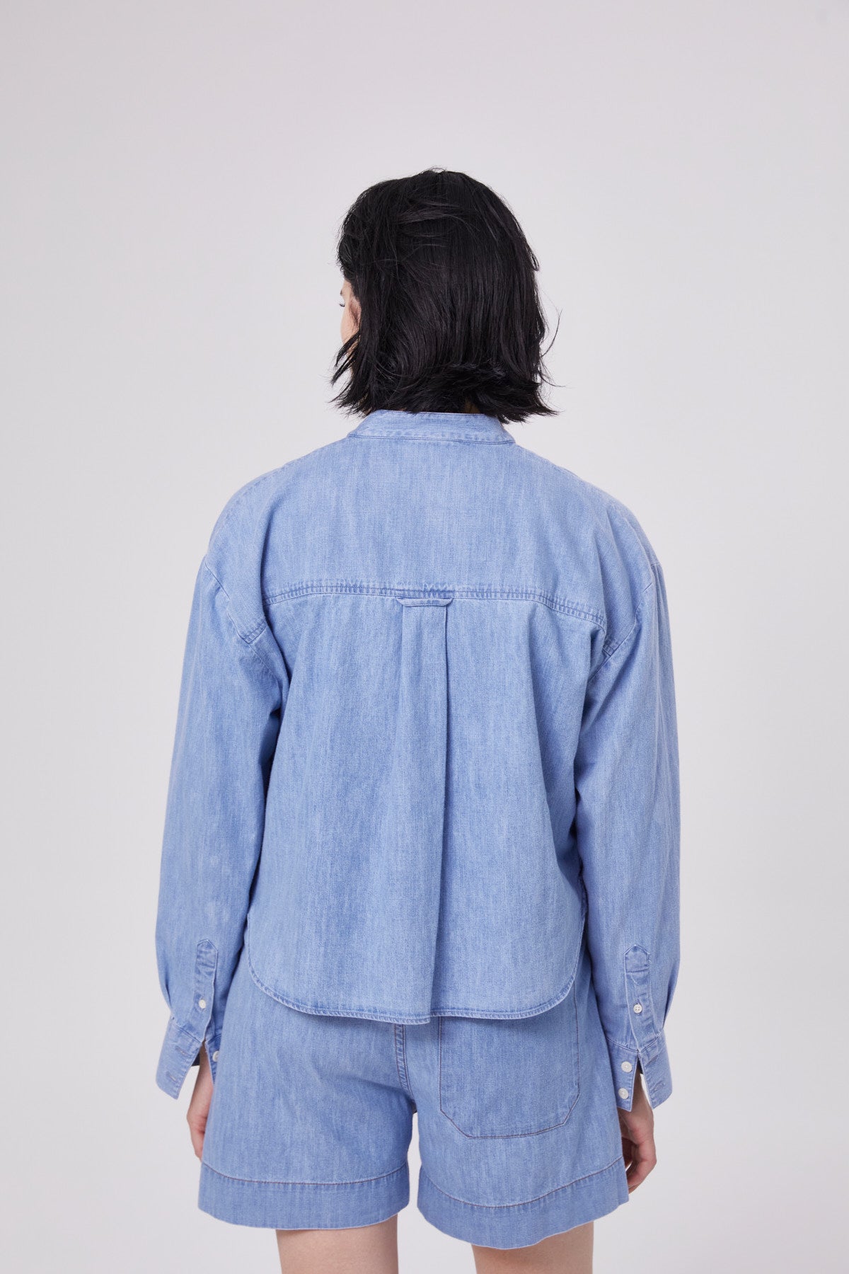 Eine Person mit kurzen schwarzen Haaren, von hinten gesehen, trägt eine hellblaue Bluse Polo Denim und passende Jeansshorts vor einem schlichten hellen Hintergrund.