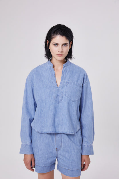 Eine Person mit kurzen dunklen Haaren trägt ein Bluse Polo Denim - ein hellblaues, langärmeliges Jeanshemd mit einer Brusttasche - und dazu passende Jeansshorts vor einem schlichten, hellen Hintergrund.