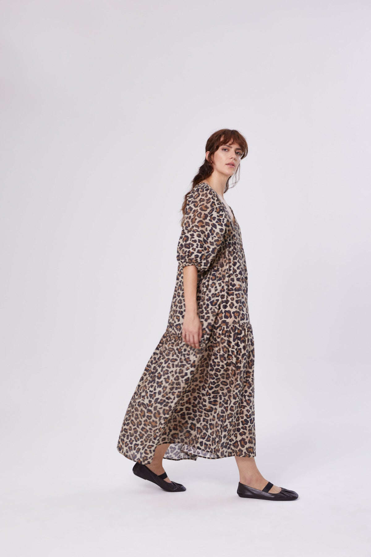 Eine Frau im Dress Fleur Leo, einem langen, locker sitzenden Kleid mit Leo-Print und schwarzen Flats, steht seitlich, der Kamera zugewandt, vor einem schlichten weißen Hintergrund.