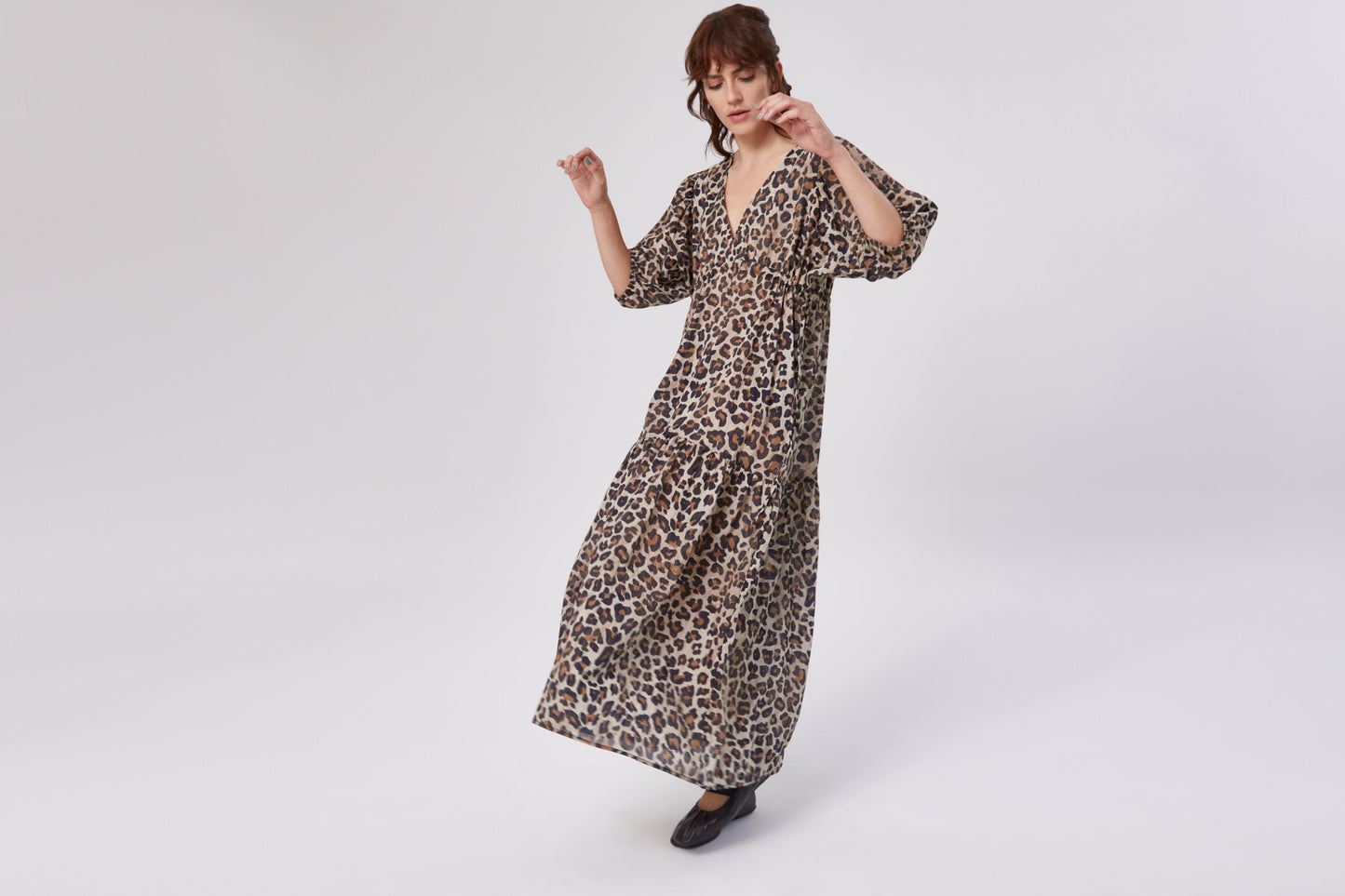 Eine Frau modelliert das Dress Fleur Leo, ein knöchellanges Kleid mit weiten Ärmeln und auffälligem Leo-Aufdruck, vor einem schlichten weißen Hintergrund. Sie trägt schwarze Schuhe und blickt leicht nach unten.