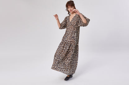 Eine Frau modelliert das Dress Fleur Leo, ein knöchellanges Kleid mit weiten Ärmeln und auffälligem Leo-Aufdruck, vor einem schlichten weißen Hintergrund. Sie trägt schwarze Schuhe und blickt leicht nach unten.