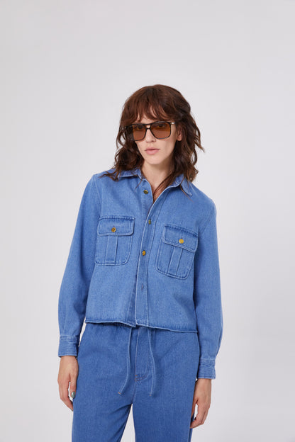 Eine Frau mit gewelltem braunem Haar trägt eine große Sonnenbrille und eine SoSUE Jacket Military aus blauem Jeansstoff mit Vordertaschen, gepaart mit einer passenden Jeanshose, vor einem schlichten hellgrauen Hintergrund.