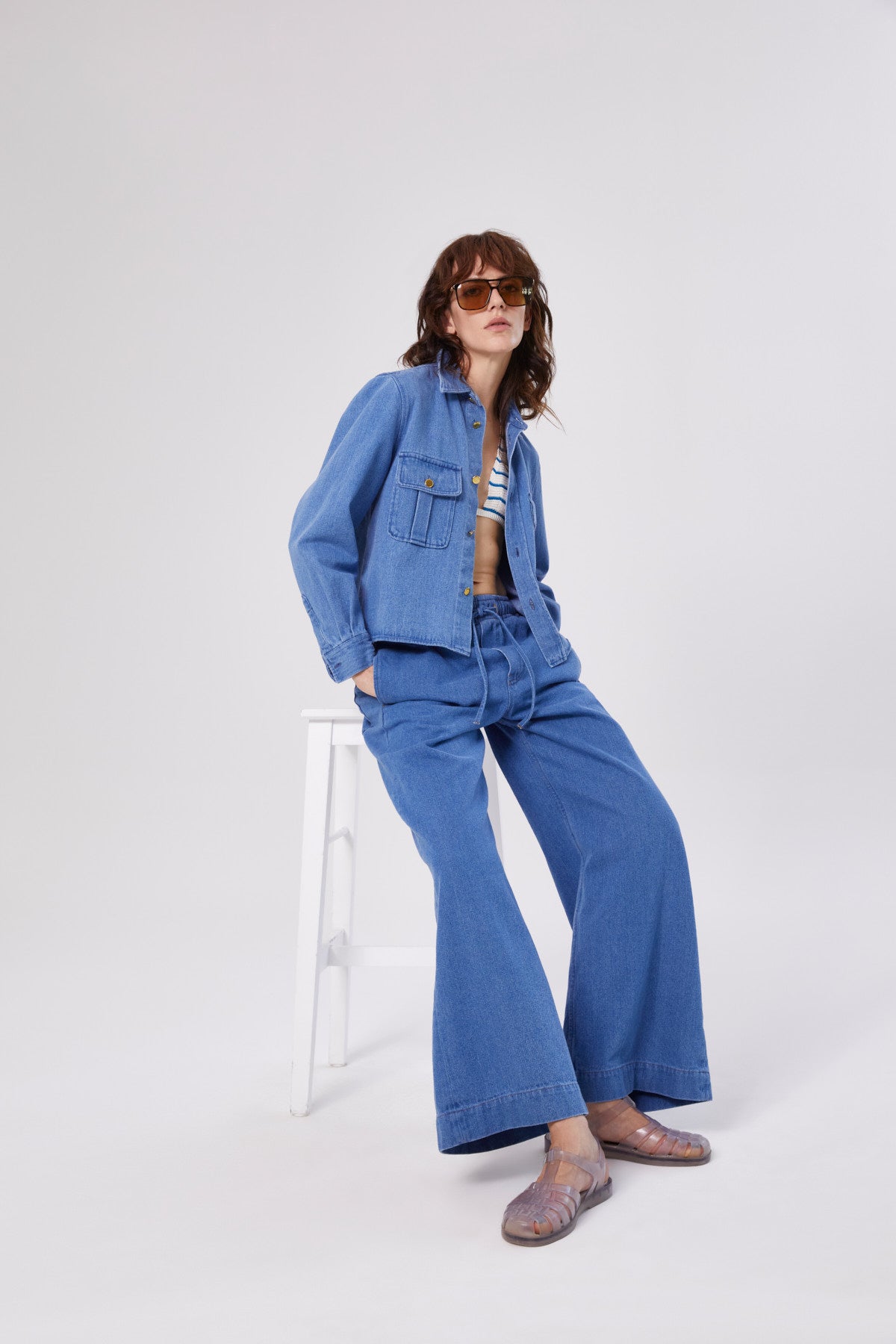 Eine Person, die eine Sonnenbrille, eine Military-Jacke, eine Jeans mit weitem Bein, ein gestreiftes Oberteil und durchsichtige Schuhe trägt, sitzt auf einem weißen Hocker vor einem schlichten hellen Hintergrund.