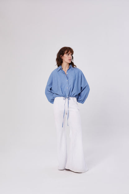 Eine Frau steht vor einem schlichten, hellen Hintergrund. Sie trägt die Bluse Antonia Drawstring Denim mit Taillenzug und weit geschnittener weißer Hose und schaut mit den Händen in den Taschen nach links.