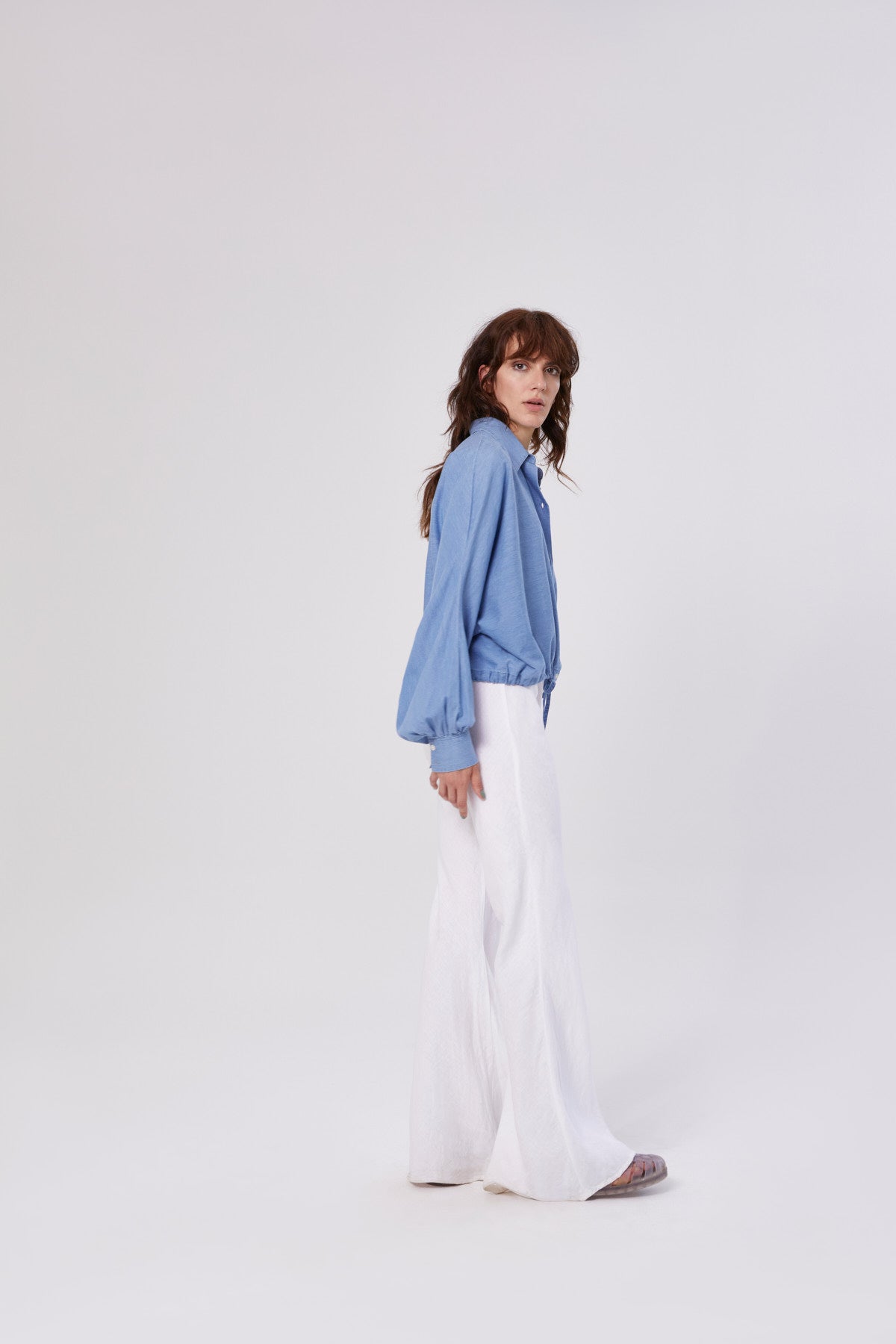 Eine Frau mit langen braunen Haaren steht seitlich vor einem schlichten weißen Hintergrund. Sie trägt die Bluse Antonia Drawstring Denim mit Ballonärmeln und einer weit geschnittenen weißen Hose.