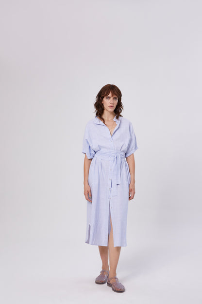 Eine Frau mit schulterlangen braunen Haaren trägt das Kleid Cowboy Button Stripes Blue White, ein hellblau-weiß gestreiftes Button-Down-Midikleid mit einer Krawatte in der Taille und durchsichtigen Schuhen, vor einem schlichten weißen Hintergrund.