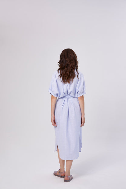 Eine Frau mit gewelltem braunem Haar blickt nach vorne. Sie trägt das Kleid Cowboy Button Stripes Blue White mit kurzen Ärmeln und einem Bindeband in der Taille, gepaart mit Sandalen, vor einem schlichten hellen Hintergrund.