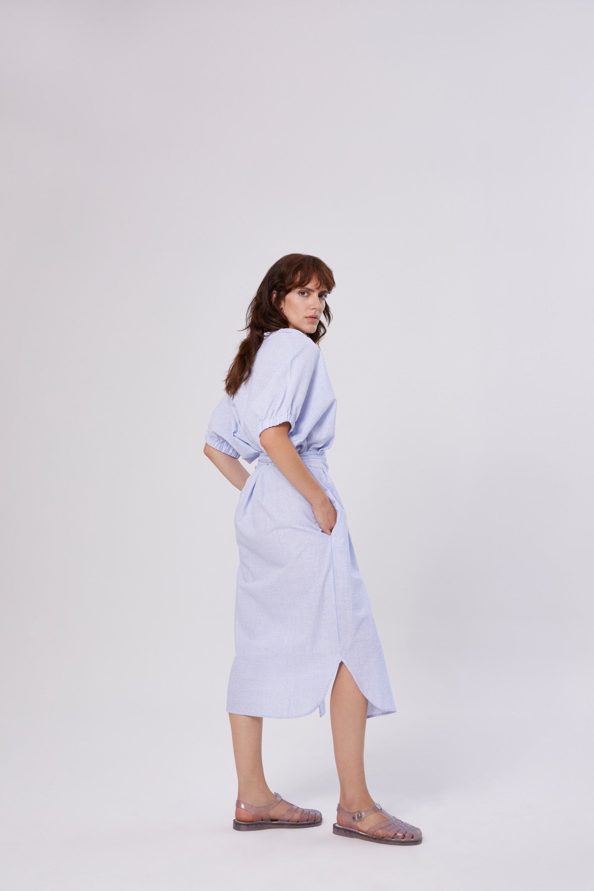 Eine Frau mit mittellangem braunem Haar steht seitlich und schaut in die Kamera. Sie trägt das Kleid Cowboy Button Stripes Blue White mit kurzen Ärmeln und durchsichtigen Sandalen vor einem schlichten weißen Hintergrund.