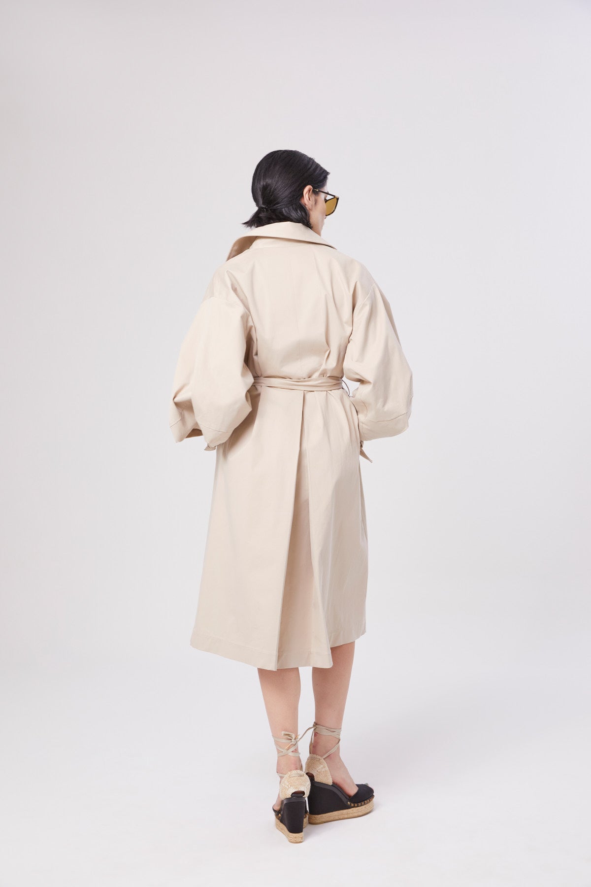 Eine Person mit dunklen Haaren in einem niedrigen Dutt steht mit dem Gesicht nach unten, trägt einen übergroßen beigen Trench Antonia mit Gürtel und schwarze Plateausandalen mit Knöchelriemen vor einem schlichten hellen Hintergrund.