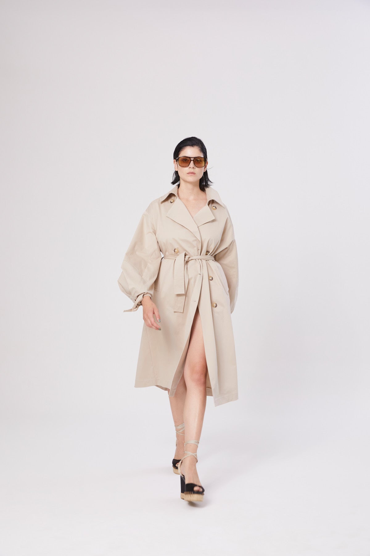Eine Frau mit dunklem Haar trägt eine Sonnenbrille und den Trench Antonia, während sie in Plateausandalen selbstbewusst nach vorne geht und vor einem schlichten weißen Hintergrund posiert.