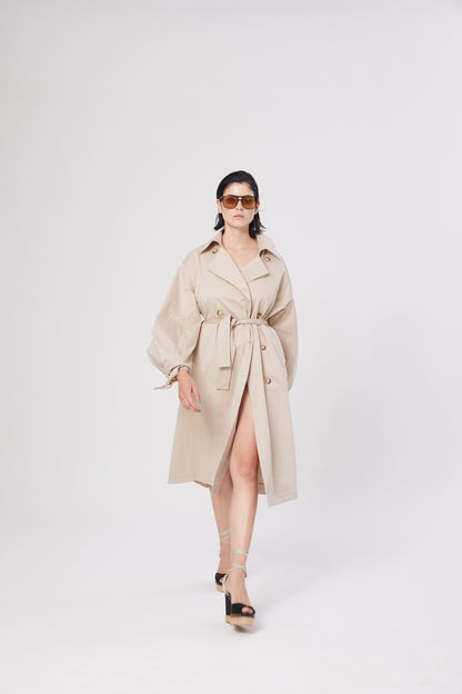 Eine Frau mit dunklem Haar trägt eine Sonnenbrille und den Trench Antonia, während sie in Plateausandalen selbstbewusst nach vorne geht und vor einem schlichten weißen Hintergrund posiert.