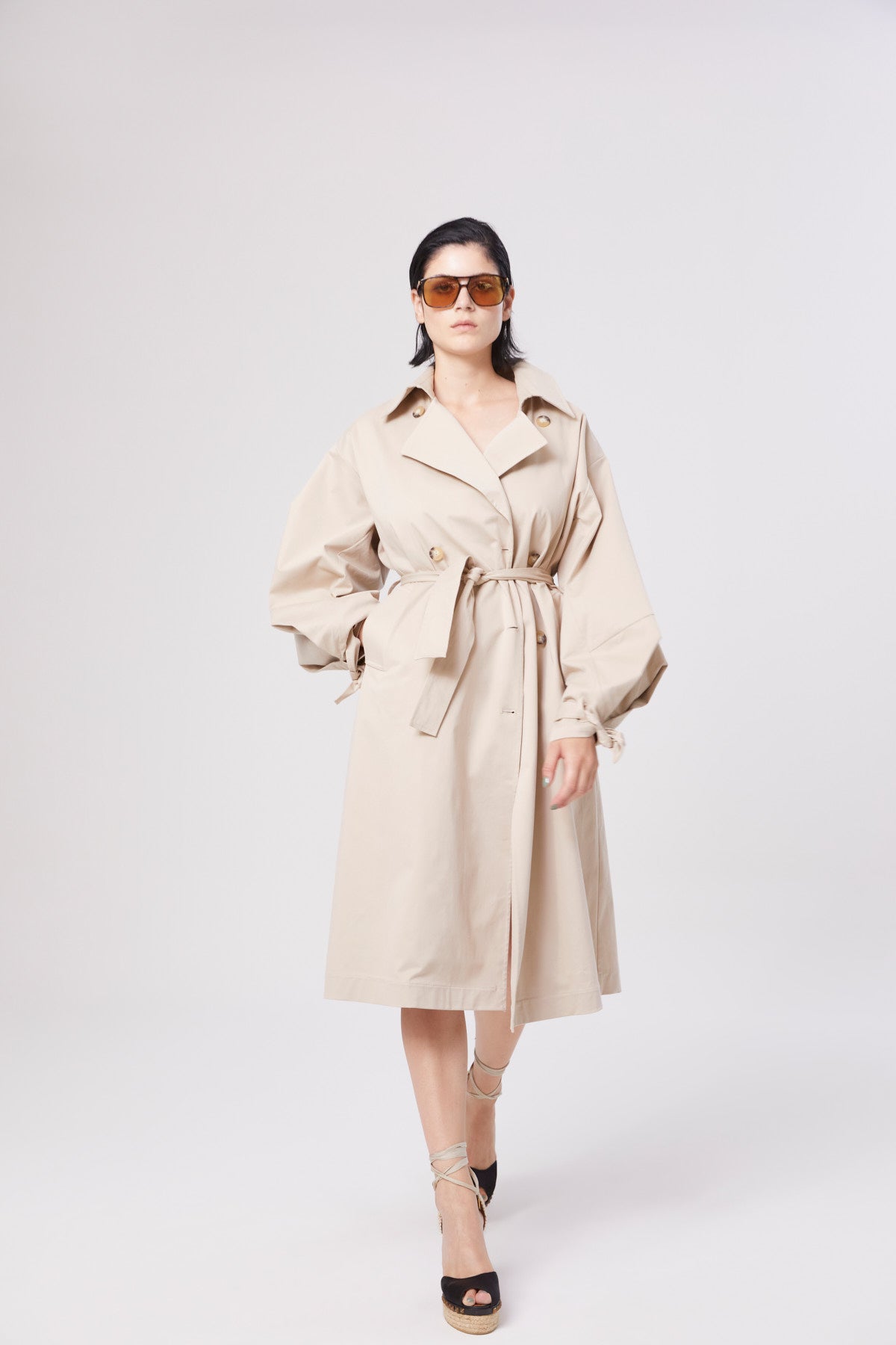 Eine Frau, die den Trench Antonia, eine Sonnenbrille und hochhackige Sandalen trägt, steht selbstbewusst vor einem schlichten weißen Hintergrund.