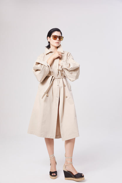 Eine Person mit Sonnenbrille und dem Trench Antonia mit Gürtel steht selbstbewusst vor einem schlichten weißen Hintergrund, die Arme leicht erhoben und die Füße in schwarzen Keilsandalen.