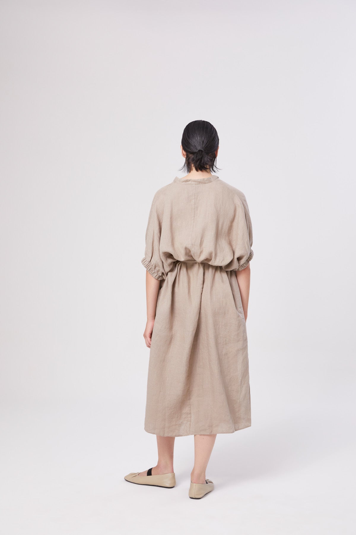 Eine Person mit kurzen dunklen Haaren blickt nach vorne und trägt das knielange, locker sitzende Kleid Cowboy Button Linen Mud mit kurzen Ärmeln und beigen Slippern. Der schlichte weiße Hintergrund unterstreicht den zeitlosen Stil dieses eleganten Kleides.