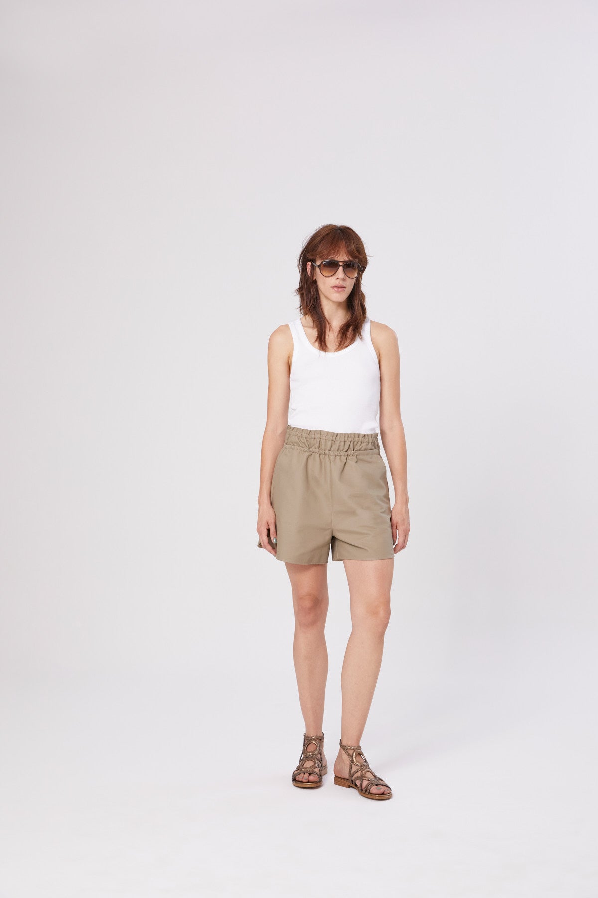 Eine Frau mit Sonnenbrille, einem weißen Tank Top Rip, khakifarbenen Shorts und braunen Sandalen steht vor einem schlichten weißen Hintergrund.