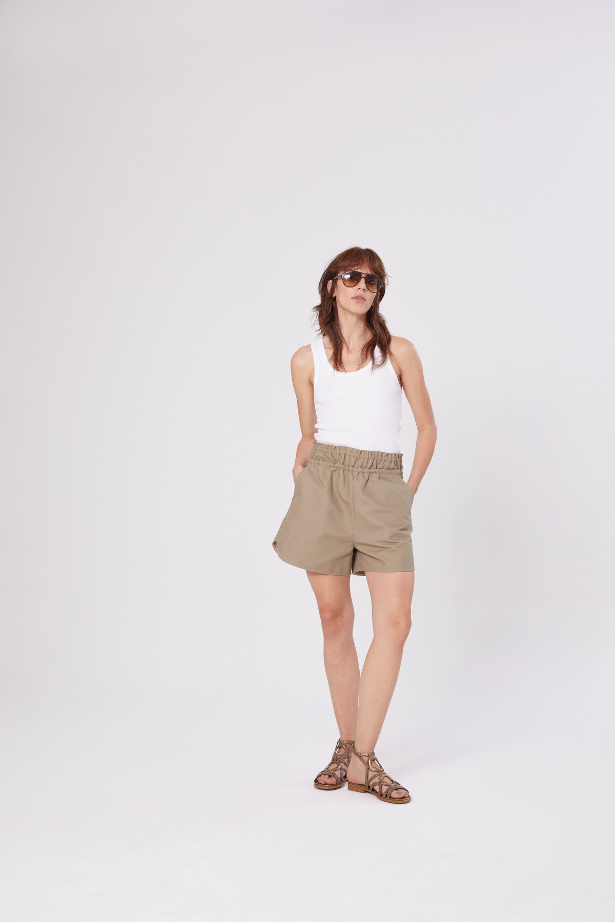 Eine Frau in einem Tank Top Rip White, khakifarbenen Shorts, braunen Sandalen und Sonnenbrille steht vor einem schlichten weißen Hintergrund und hat die Hände in den Taschen - ein lässiges Beispiel für nachhaltige Mode Damen.