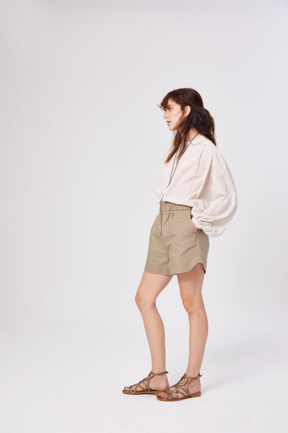 Eine Frau steht seitlich vor einem schlichten weißen Hintergrund. Sie trägt die übergroße Bluse Bella Beige mit langen Ärmeln, beigefarbene Shorts mit hoher Taille und braune Riemchensandalen.