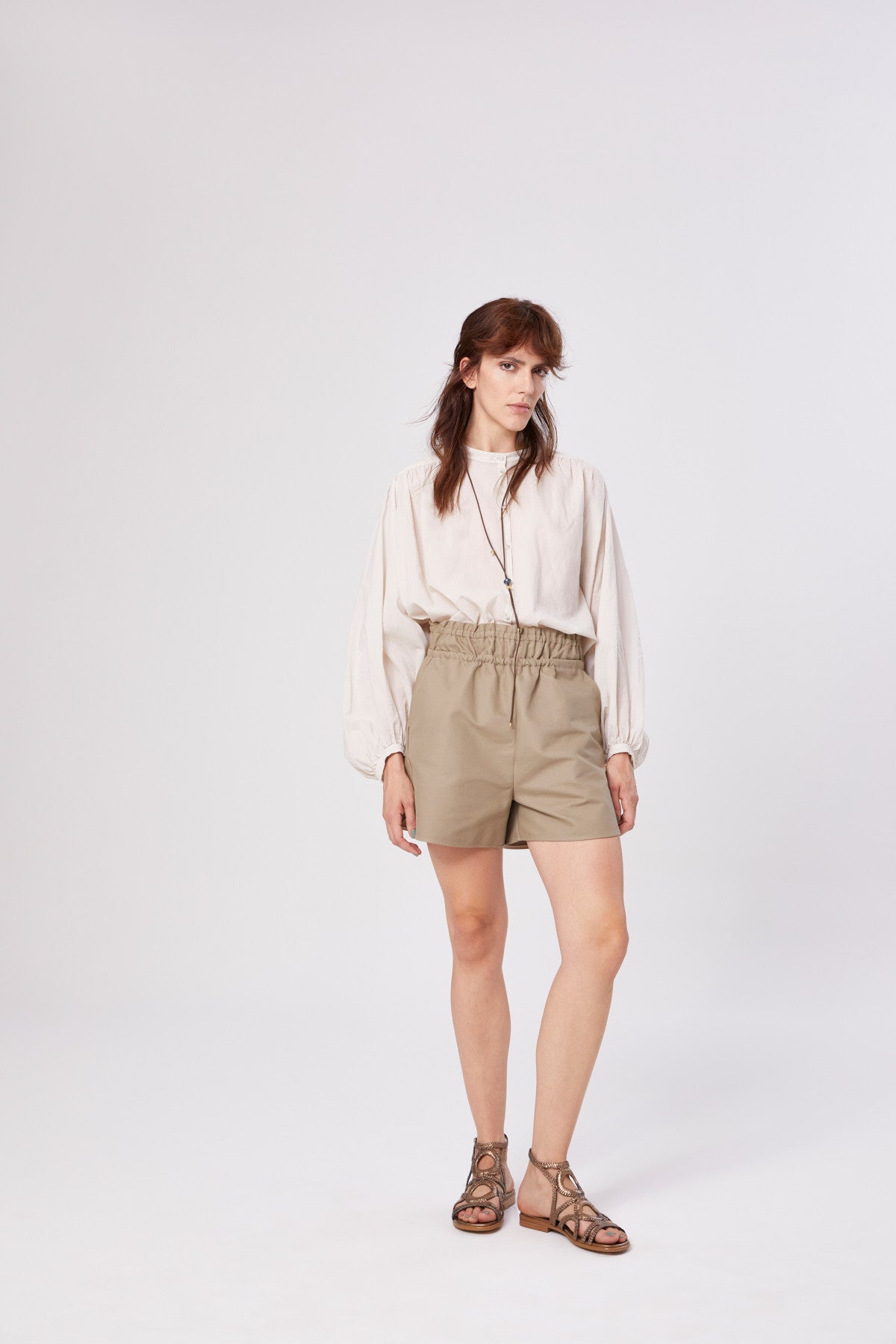 Eine Frau mit schulterlangen braunen Haaren und neutralem Gesichtsausdruck steht vor einem unifarbenen Hintergrund. Sie trägt die Bluse Bella Beige mit langen Ärmeln, helle khakifarbene Shorts mit hoher Taille und elastischem Bund sowie braune Sandalen.