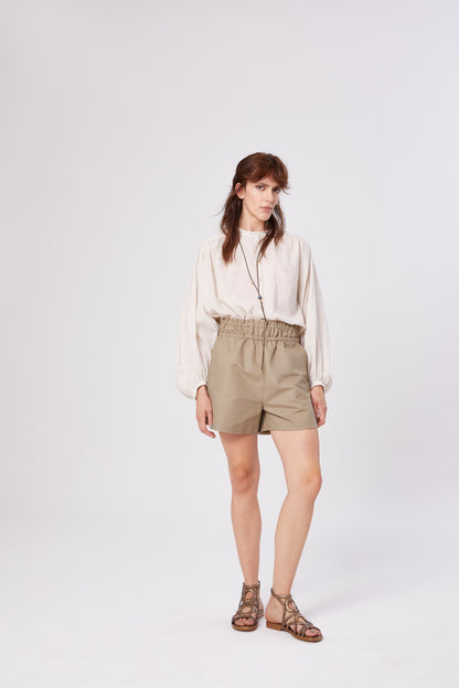 Eine Frau mit schulterlangen braunen Haaren und neutralem Gesichtsausdruck steht vor einem unifarbenen Hintergrund. Sie trägt die Bluse Bella Beige mit langen Ärmeln, helle khakifarbene Shorts mit hoher Taille und elastischem Bund sowie braune Sandalen.