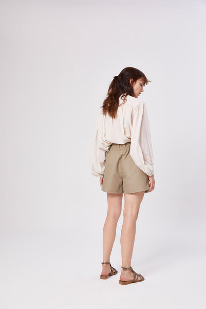 Eine Frau steht in einem neutralen Studio. Sie trägt eine Bluse Bella Beige mit langen Ärmeln, die in hoch taillierte Khaki-Shorts gesteckt wird, und braune Sandalen. Ihr lockeres, gewelltes Haar fällt ihr über den Rücken, während sie von der Kamera wegschaut.