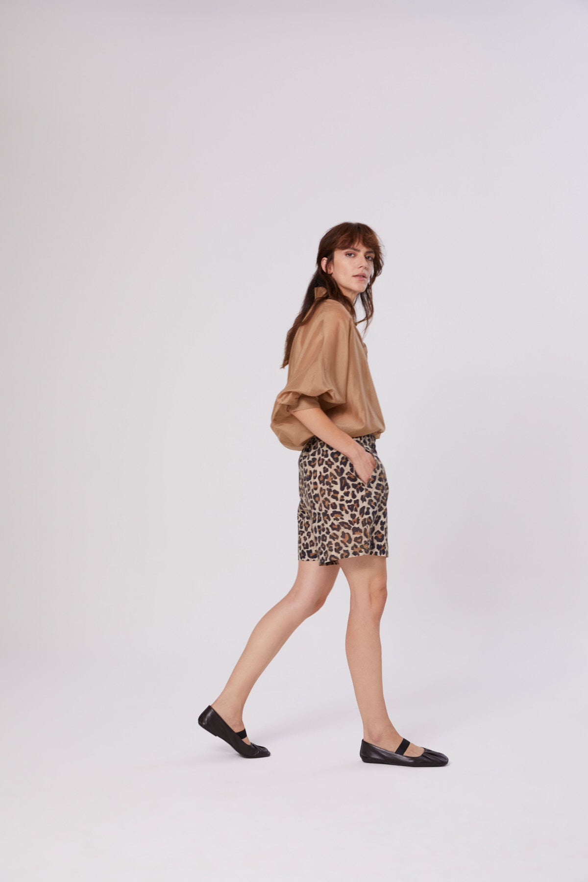 Eine Frau mit langen braunen Haaren läuft seitwärts vor einem schlichten weißen Hintergrund. Sie trägt das Bluse Antonia Short Sleeve Gold, Shorts mit Leopardenmuster und schwarze Flats.