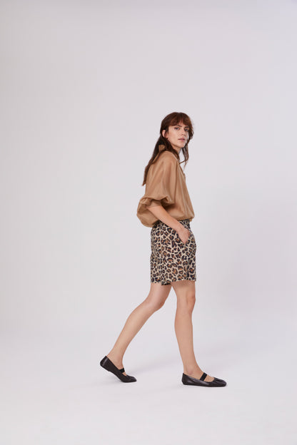 Eine Frau mit langen braunen Haaren läuft seitwärts vor einem schlichten weißen Hintergrund. Sie trägt das Bluse Antonia Short Sleeve Gold, Shorts mit Leopardenmuster und schwarze Flats.