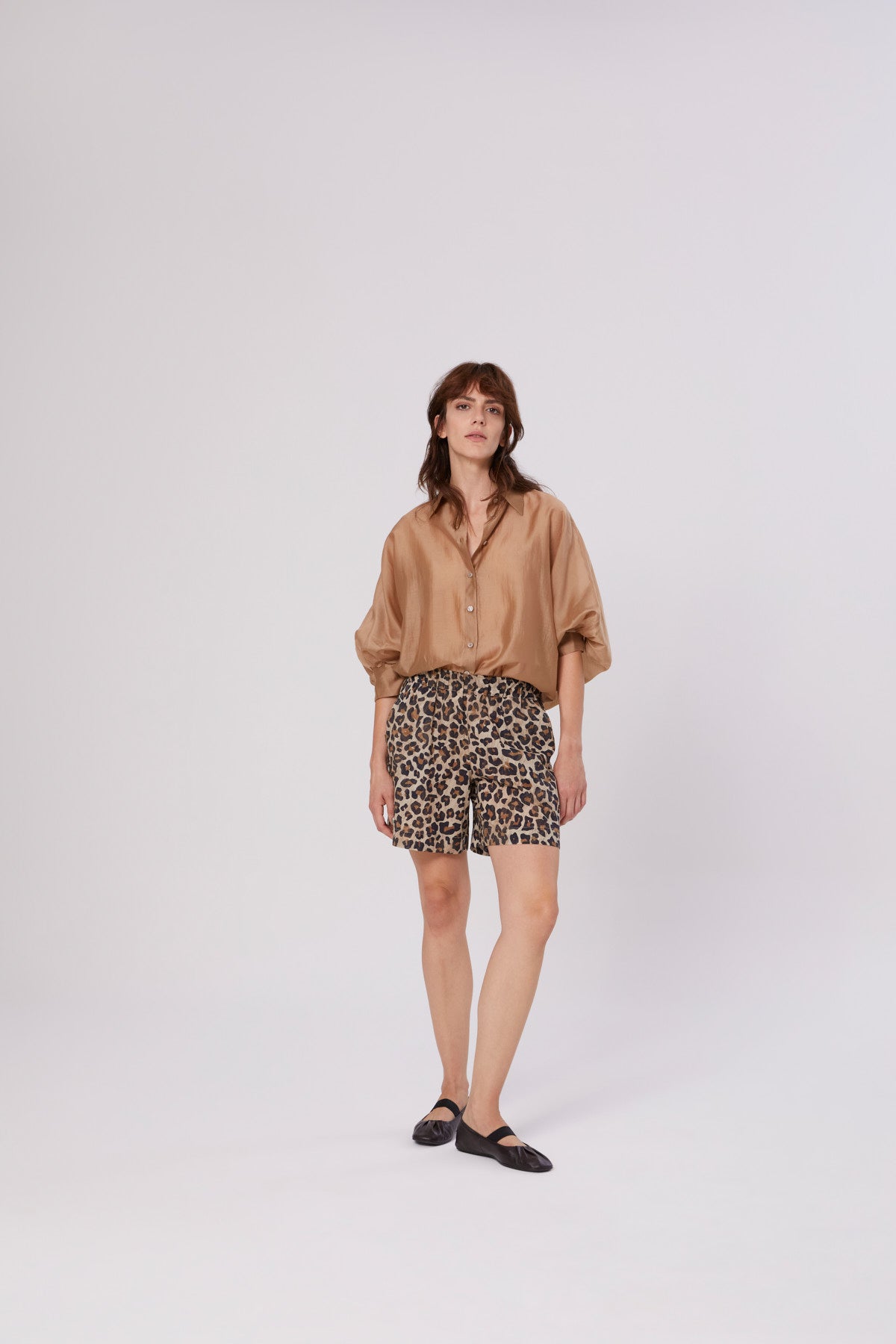 Eine Frau mit mittellangem braunem Haar und neutralem Gesichtsausdruck steht vor einem schlichten weißen Hintergrund. Sie trägt die Bluse Antonia Short Sleeve Gold, Shorts mit Leopardenmuster und schwarze flache Schuhe.