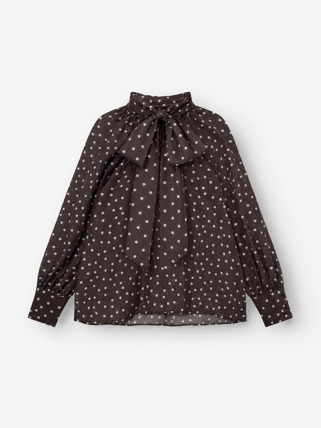 Die Bluse „Dotty Bow Brown Ecru“ ist eine langärmelige, dunkelbraune Bluse aus Baumwollvoile mit weißen Punkten, einem hohen Kragen mit einer großen Schleife am Halsausschnitt und gerafften Ärmelbündchen.