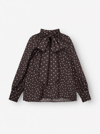 Die Bluse „Dotty Bow Brown Ecru“ ist eine langärmelige, dunkelbraune Bluse aus Baumwollvoile mit weißen Punkten, einem hohen Kragen mit einer großen Schleife am Halsausschnitt und gerafften Ärmelbündchen.