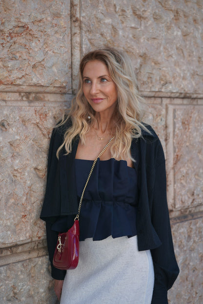 Eine Frau mit langen blonden Haaren steht vor einer strukturierten Steinmauer. Unter ihrer dunklen Jacke trägt sie ein Plissee-Top in Navy, dazu einen hellen Rock und eine rote Umhängetasche. Sie lächelt sanft und blickt leicht zur Seite.