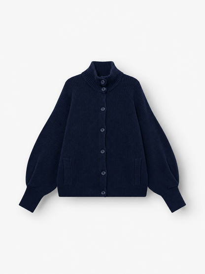 Die Strickjacke „Bomber Dunkelblau“ ist eine marineblaue Damen-Strickjacke mit langen Ärmeln, hohem Kragen, Knopfleiste vorne, gerippter Struktur, zwei Vordertaschen und leicht ausgestellten Ärmeln. Dieses stilvolle Kleidungsstück wird flach auf einem weißen Hintergrund präsentiert.